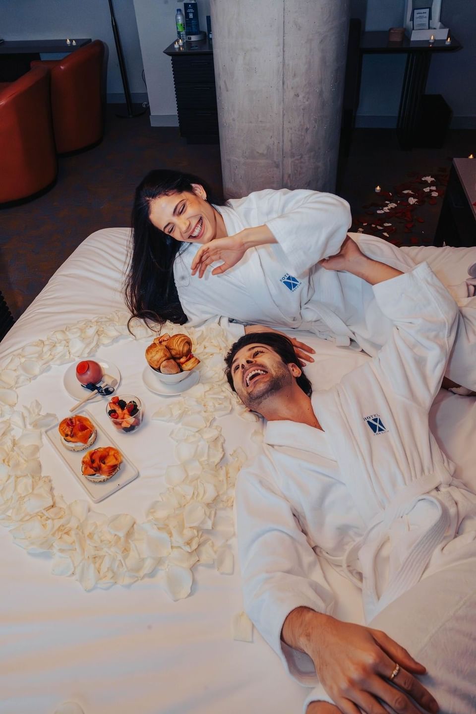 ouple laughing in robes on a bed surrounded by rose petals and pastries in a suite at Hotel X Toronto