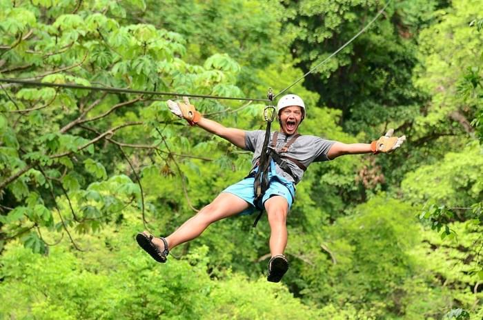 Excited man gliding on zipline over dense green forest canopy with outstretched arms near Los Altos Resort