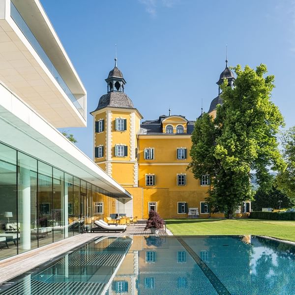 Yellow castle building next to a modern glass structure and pool, on green lawns at Falkensteiner Hotels & Residences