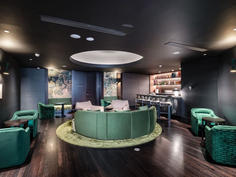 Luxurious bar lounge with green velvet seating, round tables, and a circular rug, featuring a well-stocked bar area.