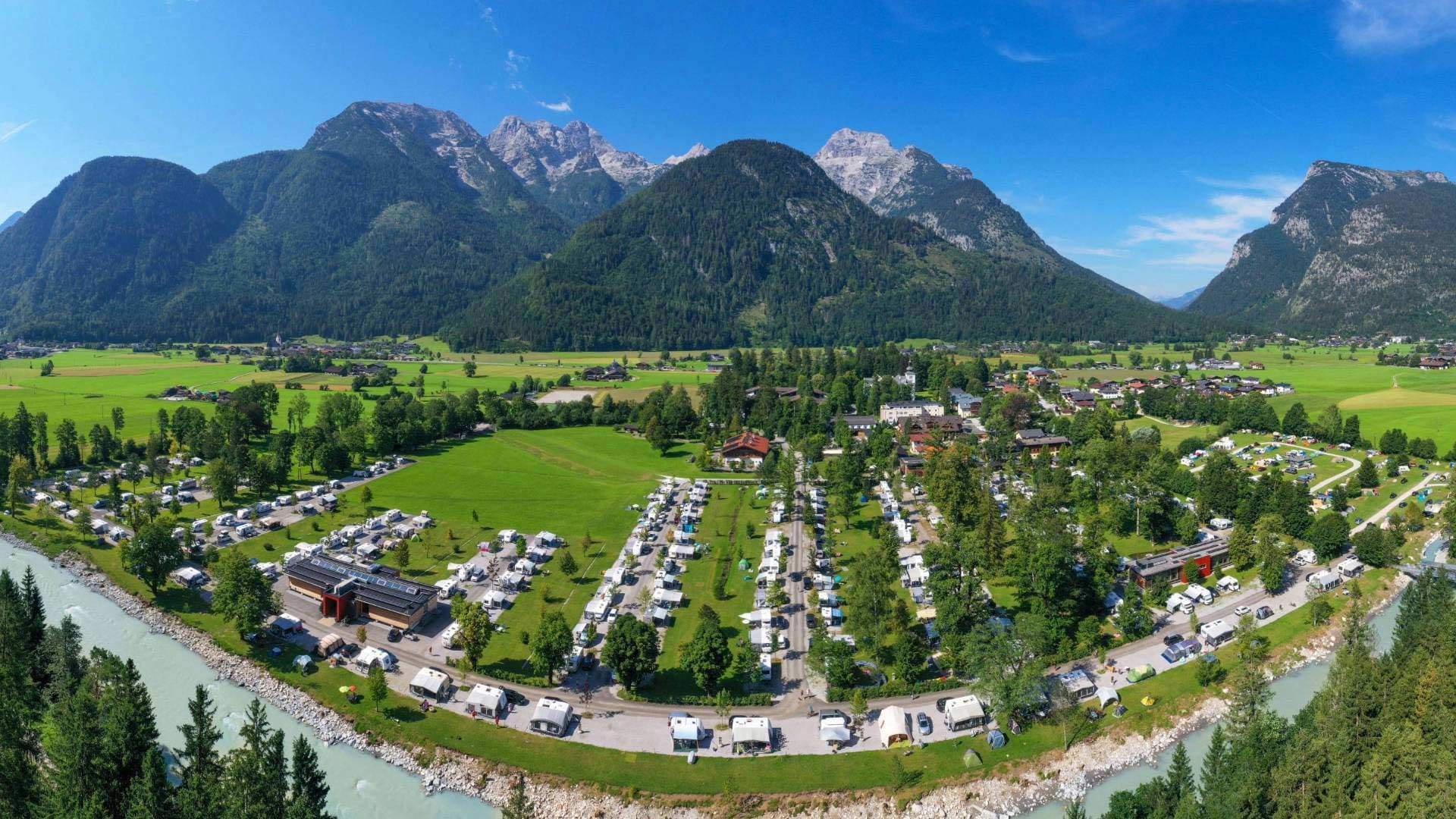 Luftaufnahme eines Campingplatzes mit vielen Wohnwagen, umgeben von Wäldern und Bergen.