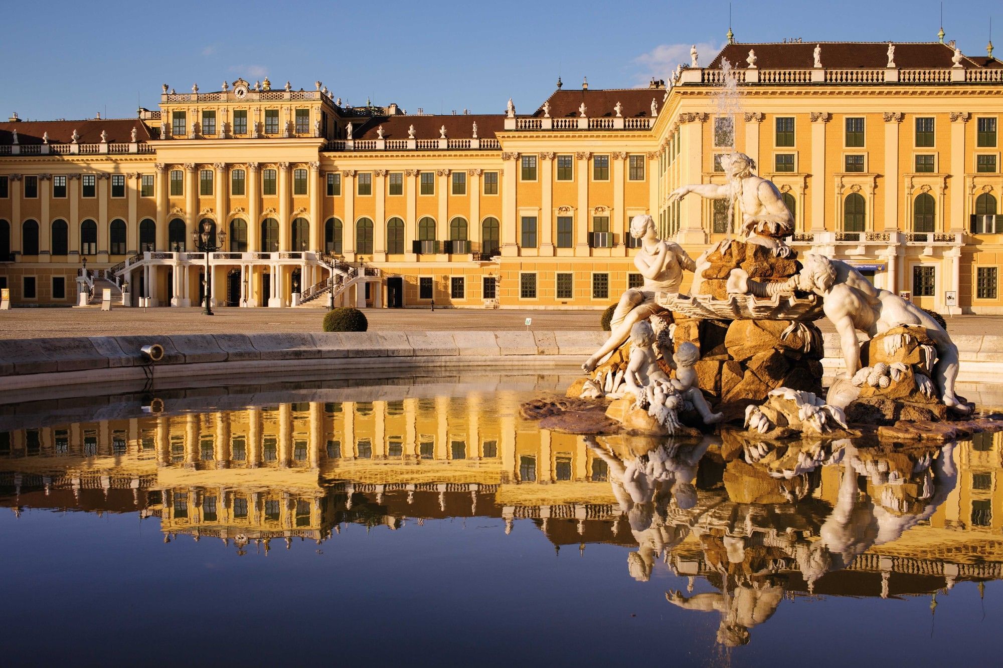 Schloss Schönbrunn in Wien mit barocker Fassade und kunstvollem Brunnen im Vordergrund, gespiegelt im Wasserbecken