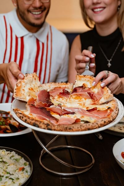 Two happy diners grabbing slices of freshly baked prosciutto pizza served on an elevated stand at Gattara