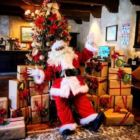 Santa Claus waving, surrounded by gifts with a decorated Christmas tree at Porta Hotel Antigua