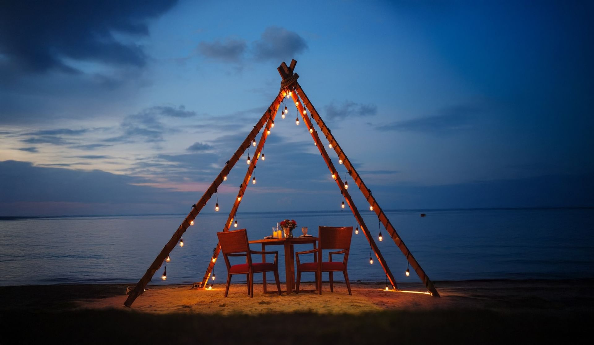 A candlelit beach dining setting under a wooden structure at dusk
