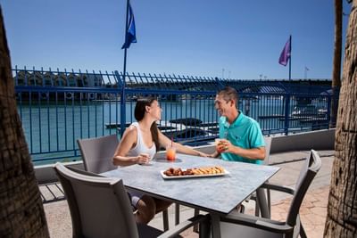 Couple enjoying drinks and appetizers at waterfront patio table with blue bridge view at London Bridge Resort