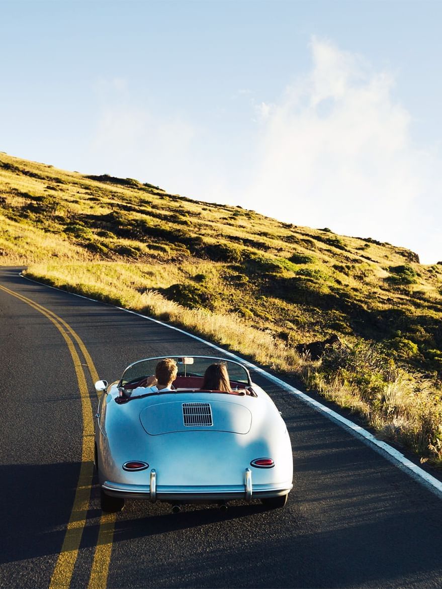 Silver convertible driving in a lane by a grassy hillside under a blue sky near Warwick Grand Place Brussels