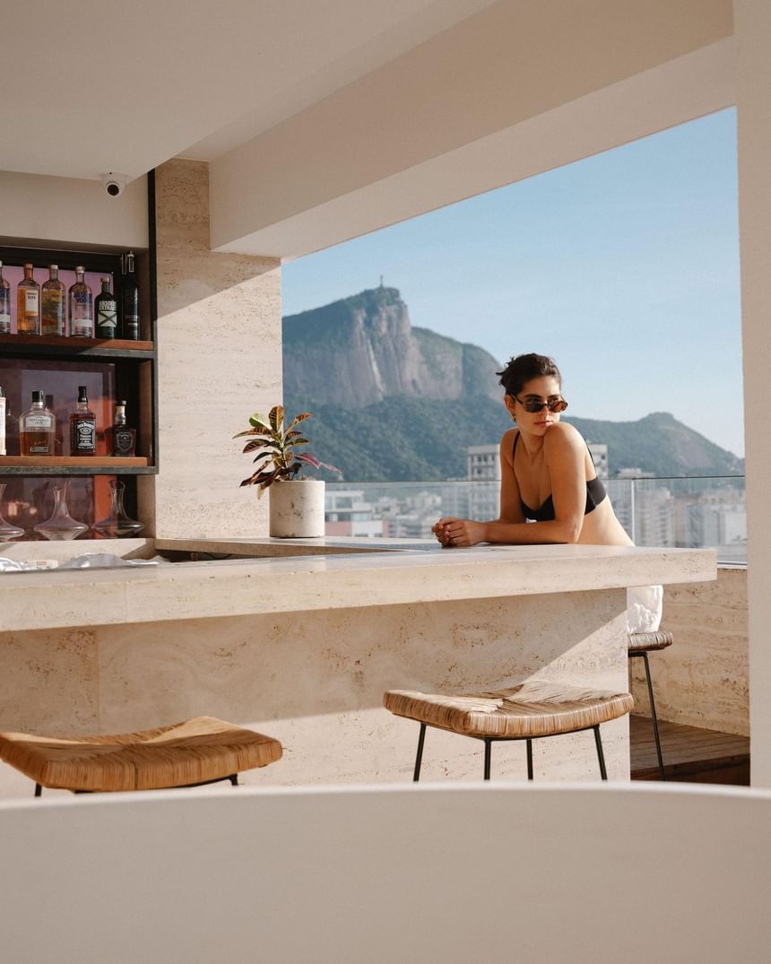 Terraço do Janeiro Hotel com um bar com vista do RJ e uma mulher sentada a espera de um drink