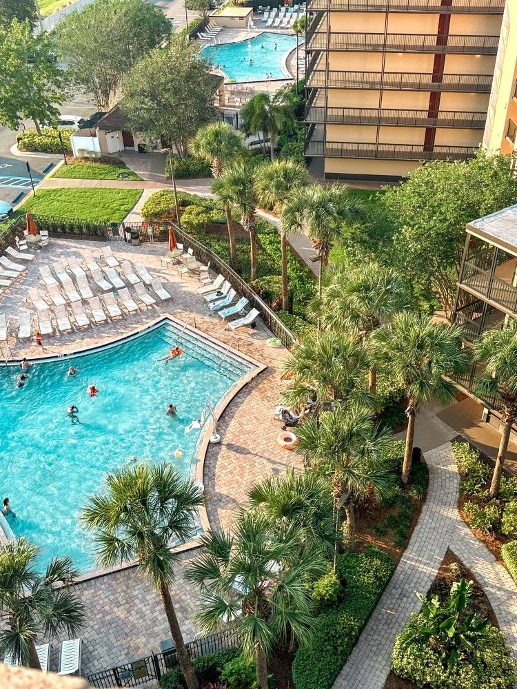 An aerial shot of a sparkling blue swimming pool with people in it surrounded by lounge chairs and palm trees. 