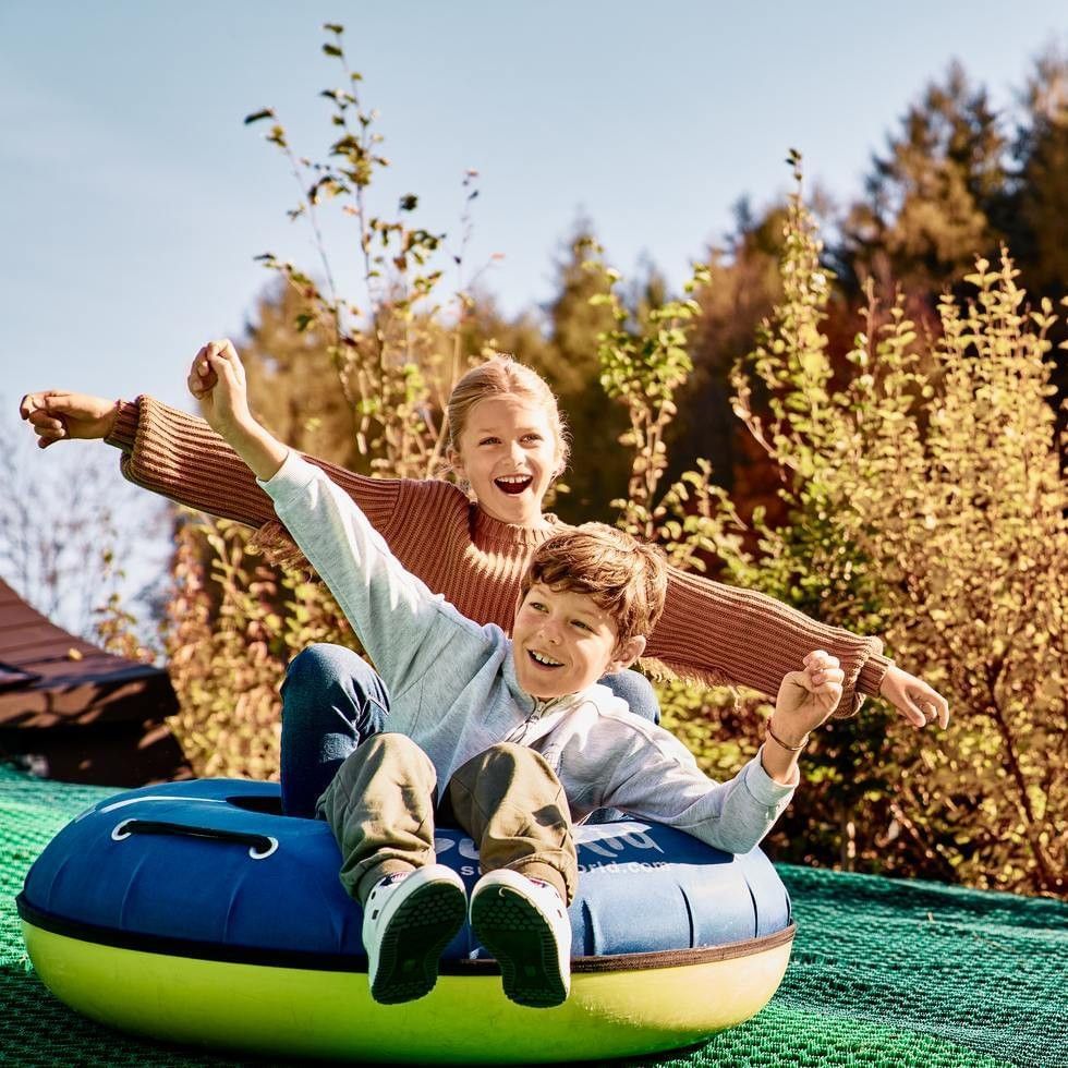 Two kids laughing on a blue tube slide, surrounded by a golden autumn forest near Falkensteiner Hotels & Residences