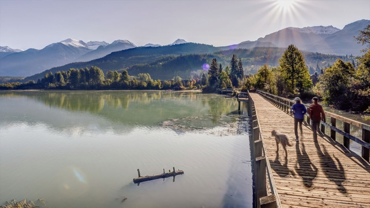 Two people and a dog walk over a wooden bridge near a lake with mountains in the background.
