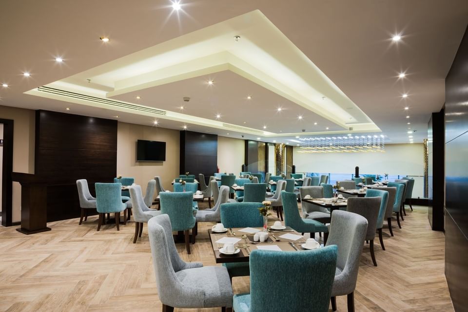 Blue armchairs by square dining tables under a recessed ceiling in Naviti Restaurant at Naviti By Warwick Dammam