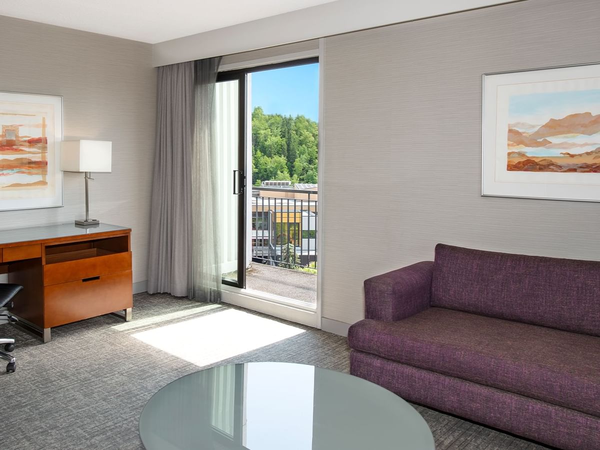 A hotel suite living room with couch, table, desk, chair, framed art, and balcony with view.