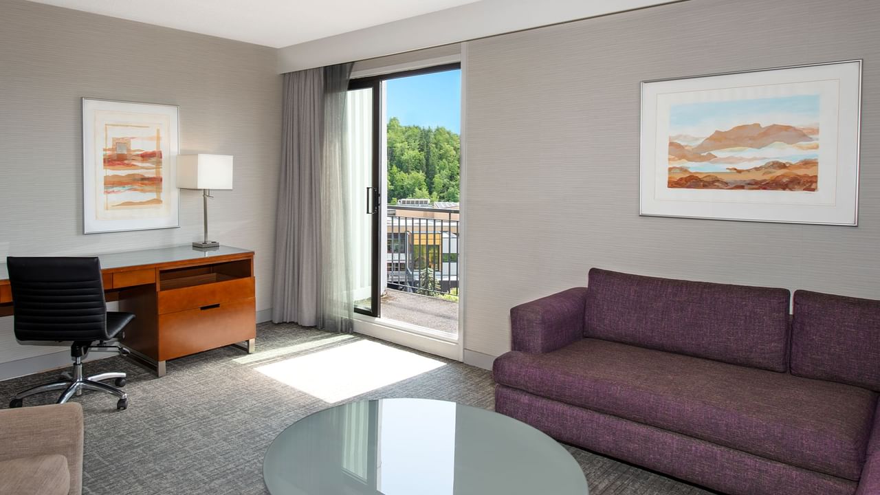 A hotel suite living room with couch, table, desk, chair, framed art, and balcony with view.