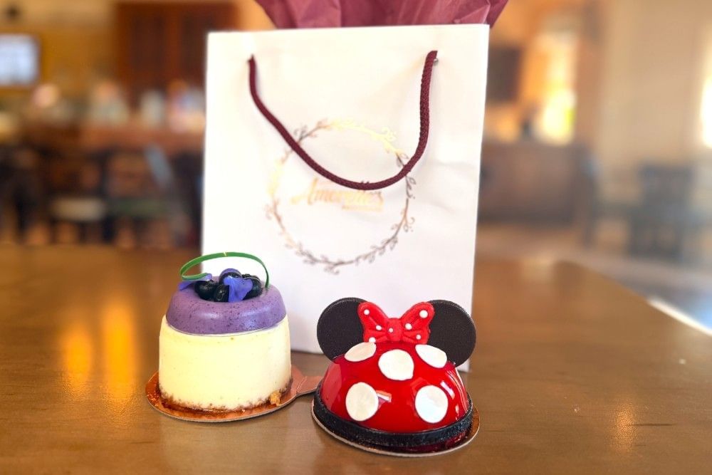A close-up of two beautiful desserts, one designed to look like Minnie Mouse with red and white polka dots, sitting on a table beside a white gift bag with the Amorette's logo from Disney Springs.