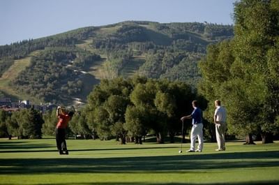 Golfer swinging on a green field with two spectators and hills behind at Hotel Park City Autograph Collection