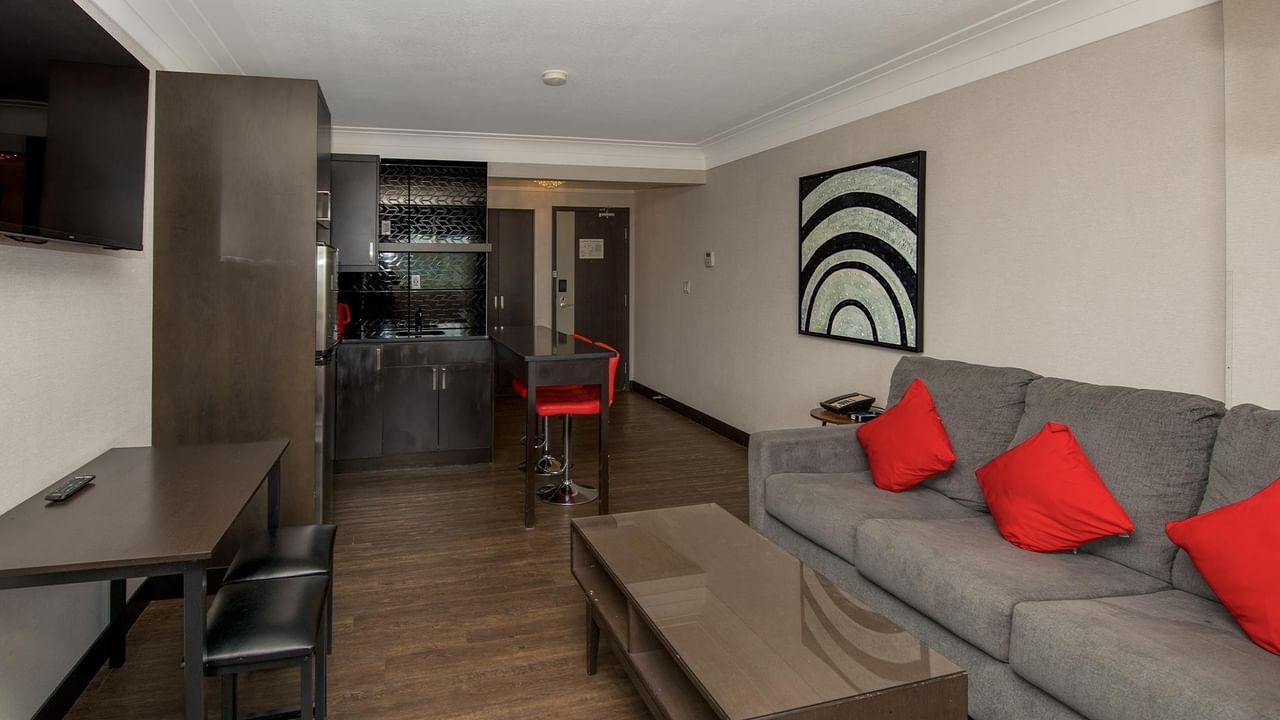 Modern hotel suite with gray sofa, red accent pillows, kitchenette, and dining area with wood-style flooring.