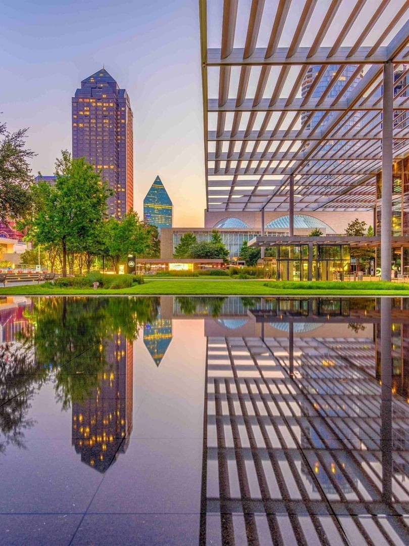 Modern city skyline by a park under a metal canopy surrounding a reflecting pool at Warwick Melrose Dallas
