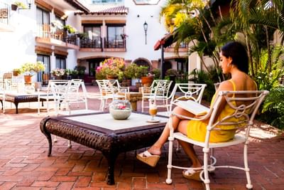 Woman in yellow dress reading a book at an outdoor patio with white chairs and brick floor.