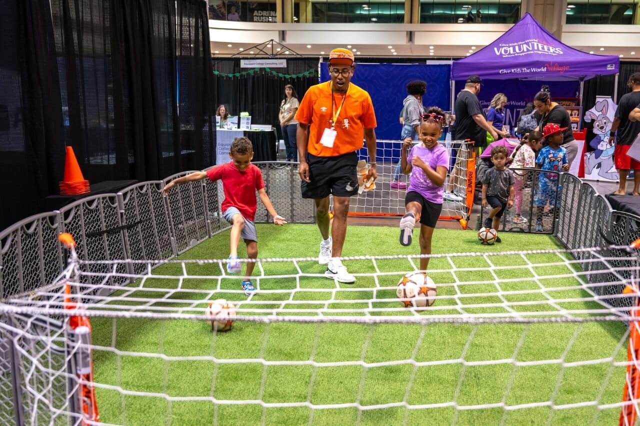 A man with an orange t-shirt and hat stands between two kids kicking soccer balls into an indoor soccer net.