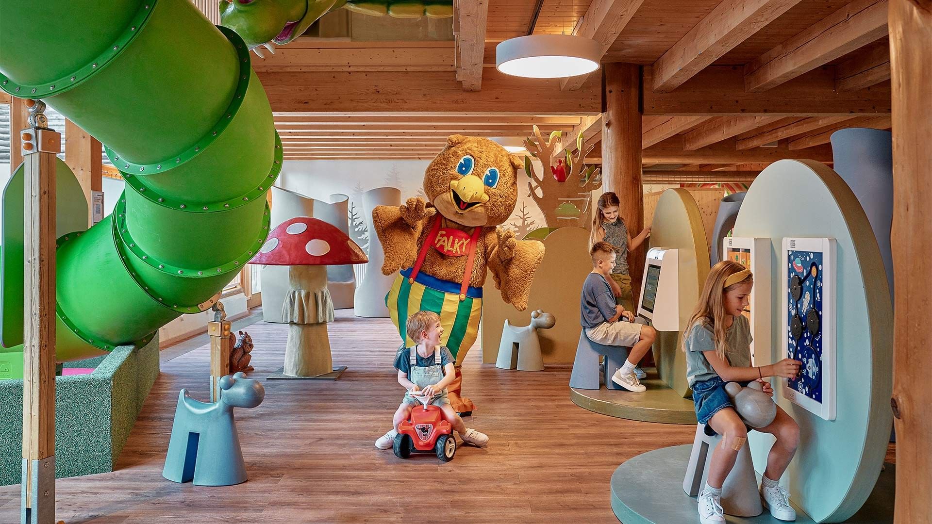 Kinder spielen in einem Raum mit Holzboden und einer Falk-Maskottchen-Statue.
