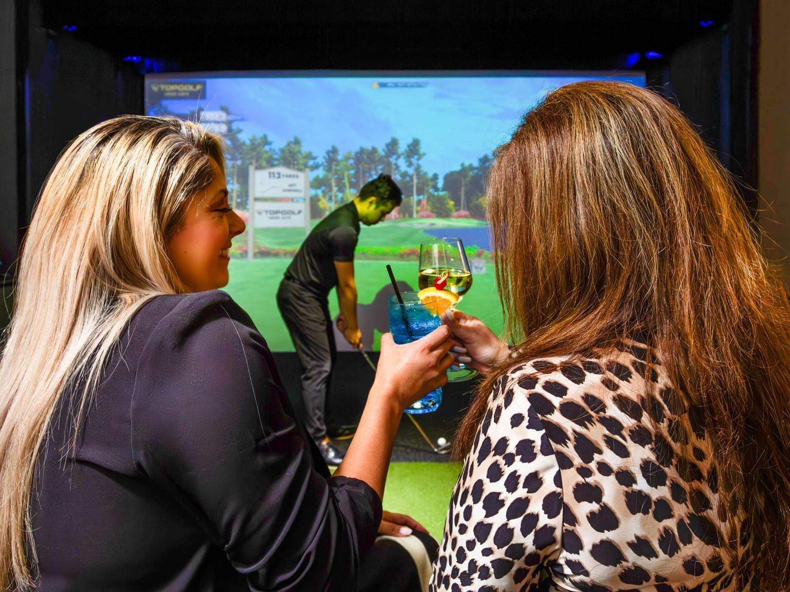 two people in foreground with drinks to cheers and man playing golf simulator in background