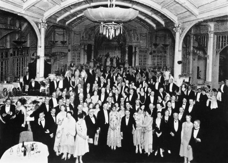 A large group of formally dressed people gathered in a grand hall, some seated and others standing.