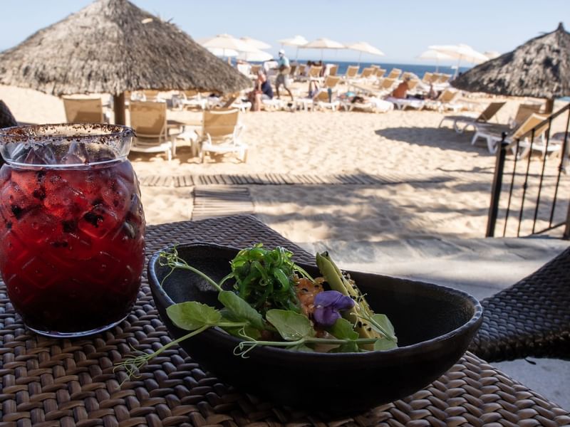 Red drink in a glass with a salt rim next to a small dish of salad on a table by the beach at Hacienda Del Mar Los Cabos