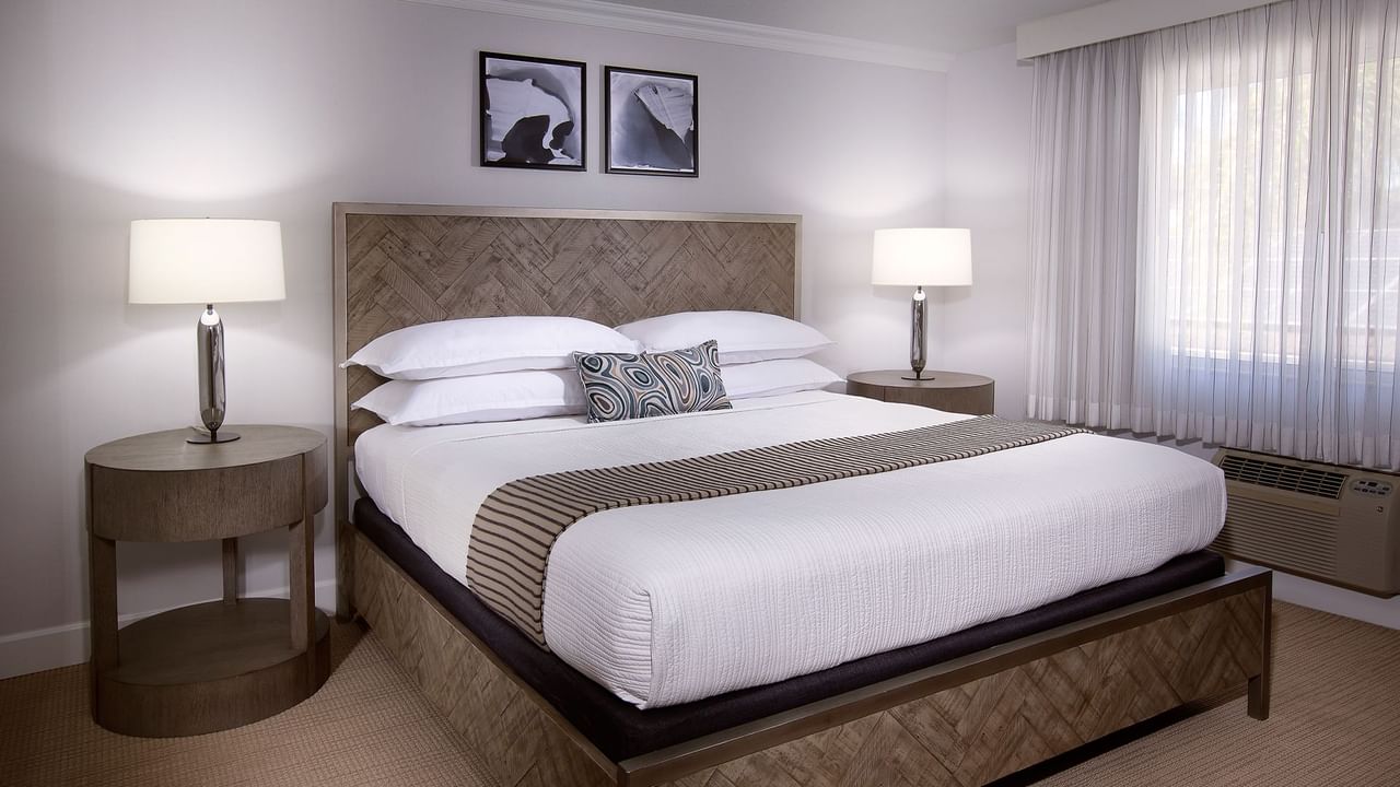 A bedroom with a large bed, two nightstands with lamps, framed pictures, and a window with curtains.