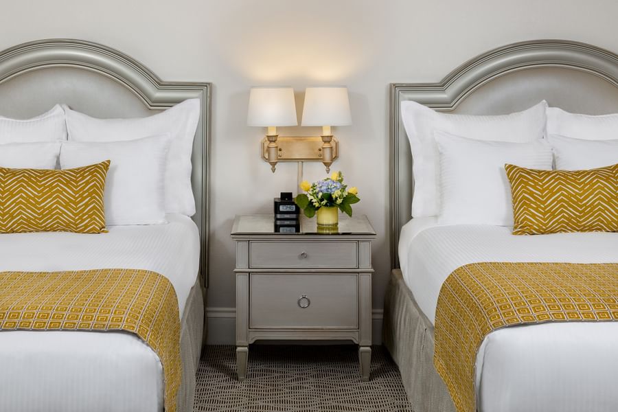 Grey nightstand with flowers by wall sconces under two bed headboards in Classic Double Accessible at Warwick Melrose Dallas