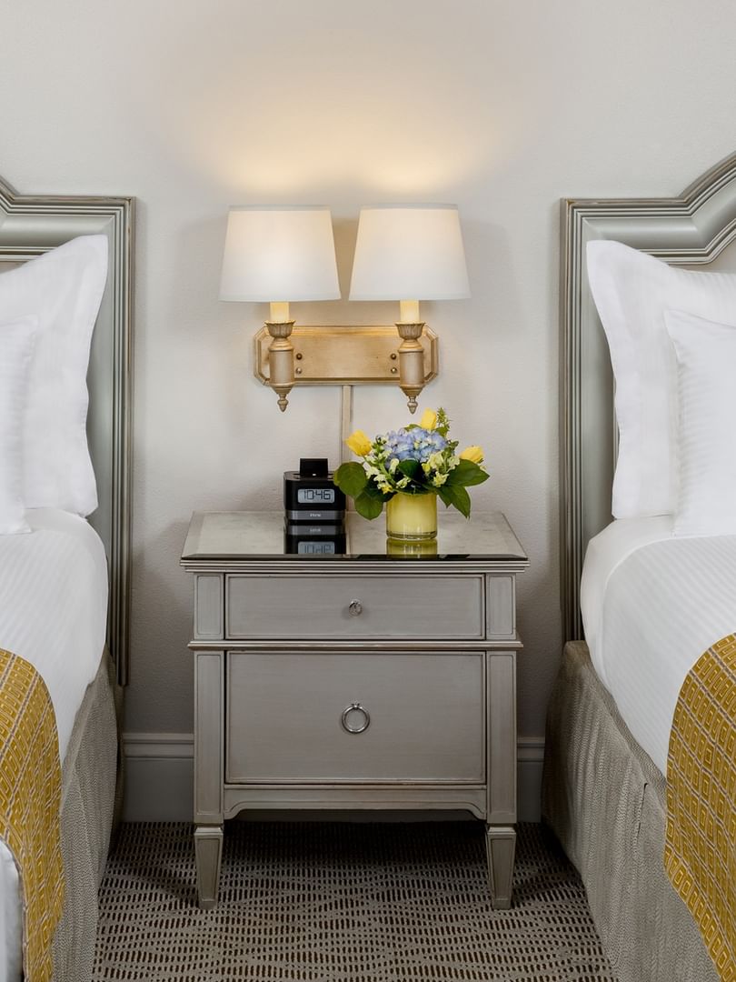 Grey nightstand with flowers by wall sconces under two bed headboards in Classic Double Accessible at Warwick Melrose Dallas