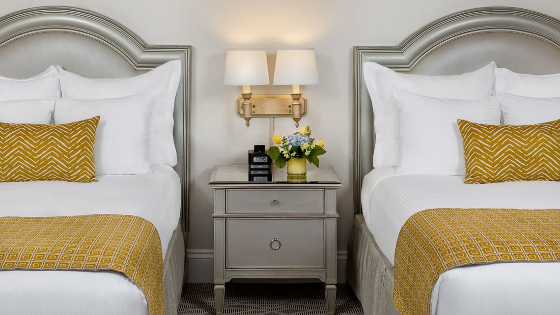 Grey nightstand with flowers by wall sconces under two bed headboards in Classic Double Accessible at Warwick Melrose Dallas