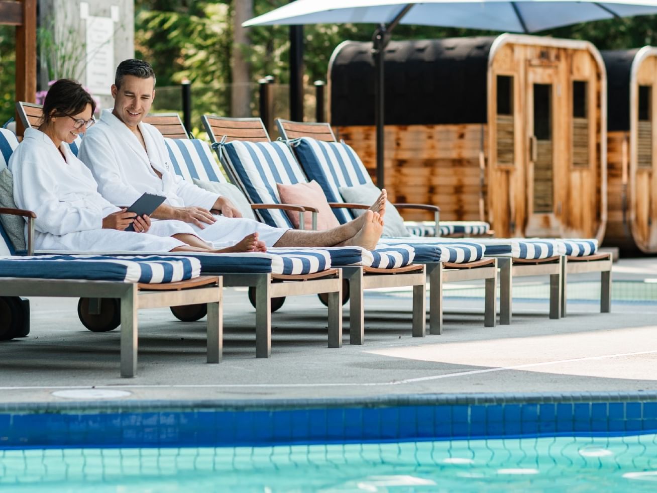 Couple relaxing on lounge chairs by a pool with wooden saunas in the background.