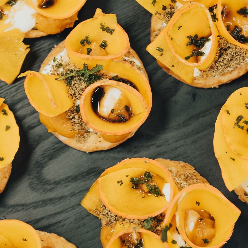 Close-up of six small dishes topped with orange-colored slices and garnish, on a dark surface.