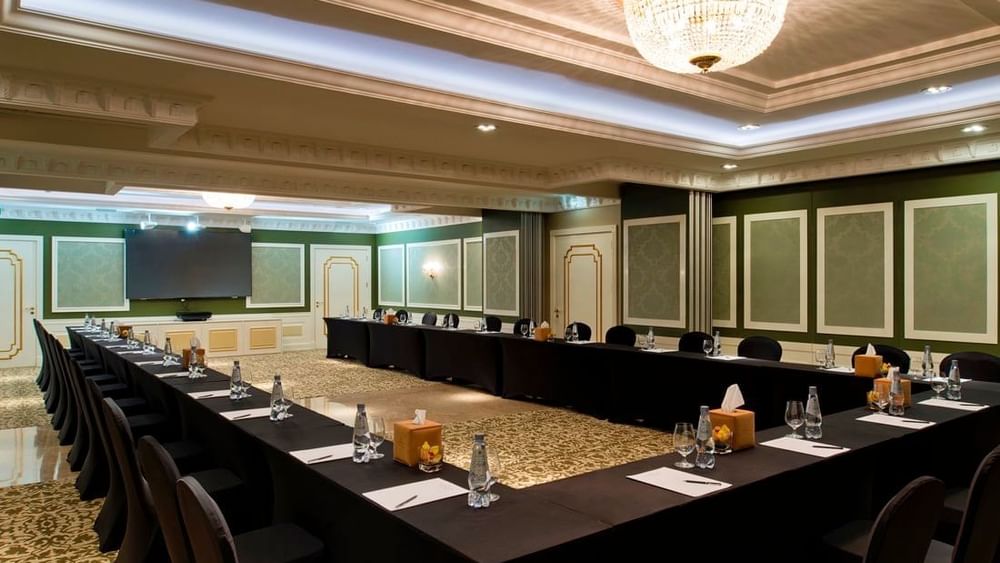 U-shaped meeting table with black chairs by a screen under a tiered ceiling in Emerald Room at Warwick Doha Hotel