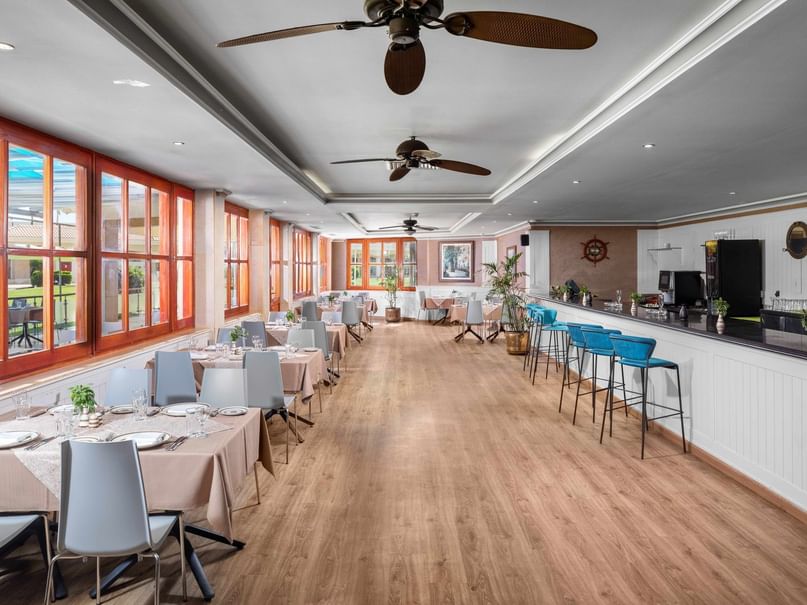 A restaurant with wooden floors, ceiling fans, tables, chairs, a bar, and large windows.