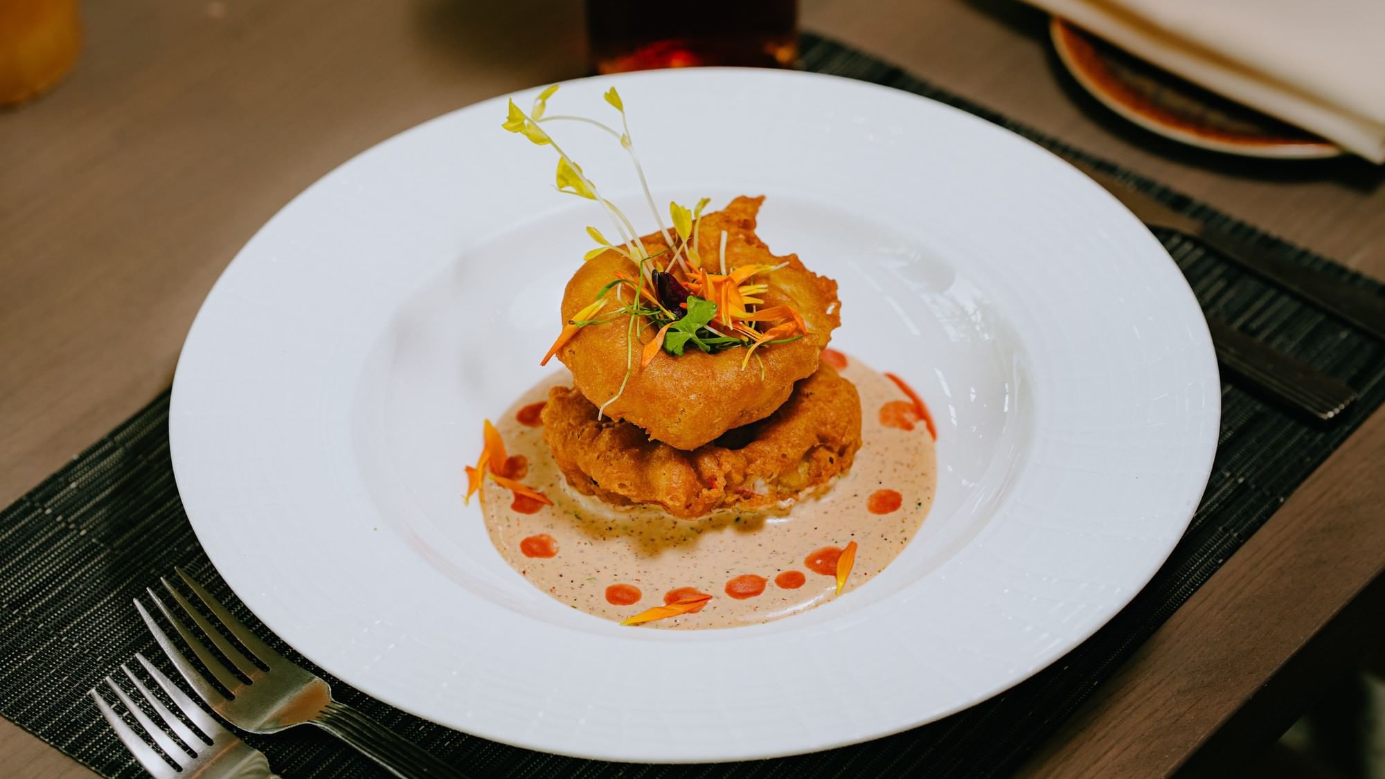 A plate with two fried items topped with garnish and sauce, accompanied by forks on a placemat.