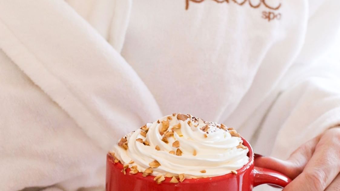 Mug filled with a creamy, whipped-cream-topped beverage, held by girl in a white pause spa robe at Paramount Hotel Midtown