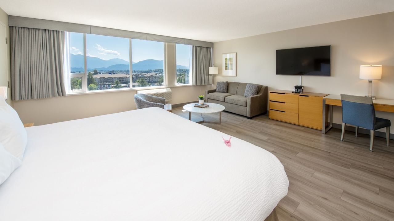 A hotel room with a bed, desk, couch, chairs, and a large window overlooking mountains.