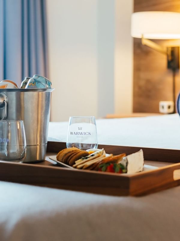 Breakfast tray with cheese and crackers by an ice bucket on a bed in In-Room Dining at Warwick Denver