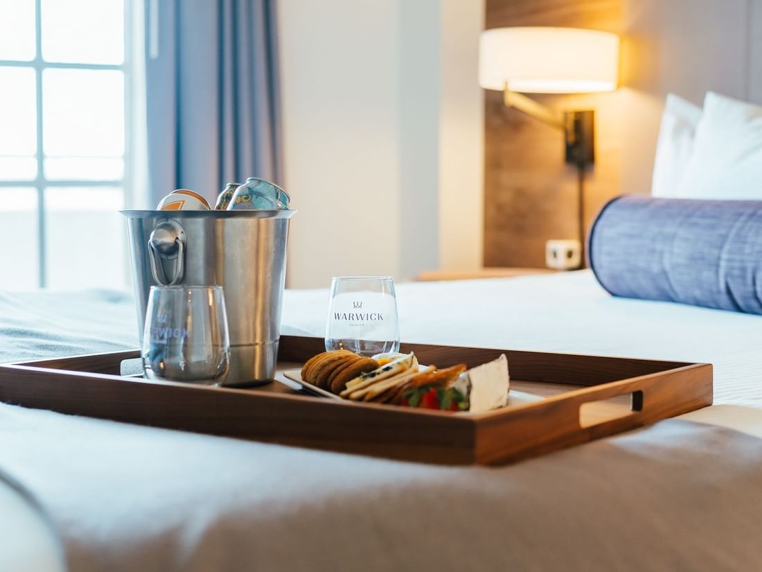 Breakfast tray with cheese and crackers by an ice bucket on a bed in In-Room Dining at Warwick Denver