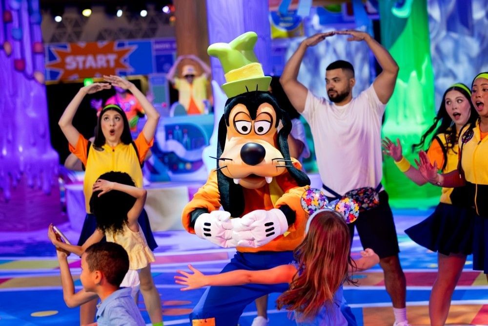 Goofy dances with arms outstretched alongside several adults and children at a dance party.