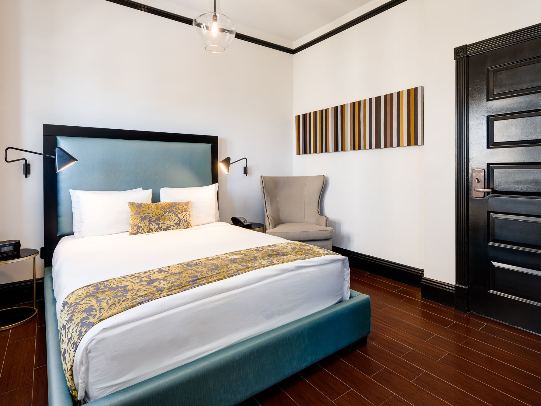 A hotel room with a queen bed, bedside lamps, chair, striped artwork, and a black door.