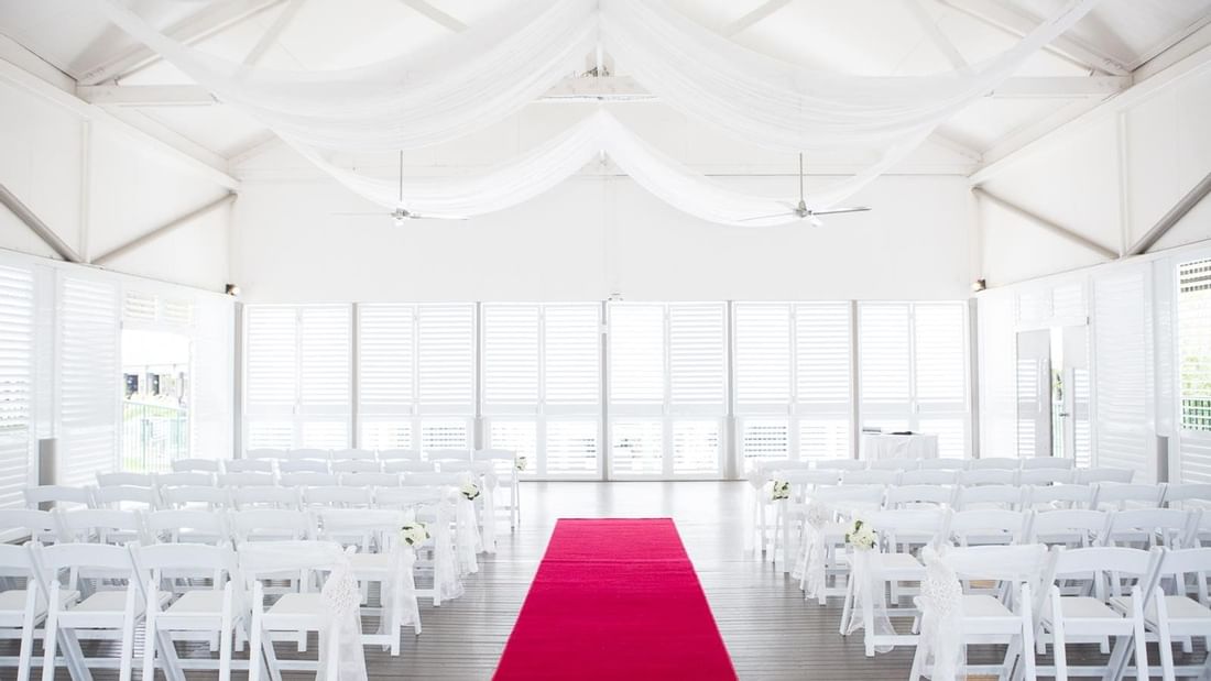 An elegant wedding ceremony setup featuring white chairs, a red aisle carpet, and draped fabric in Mercure Hotel Townsville