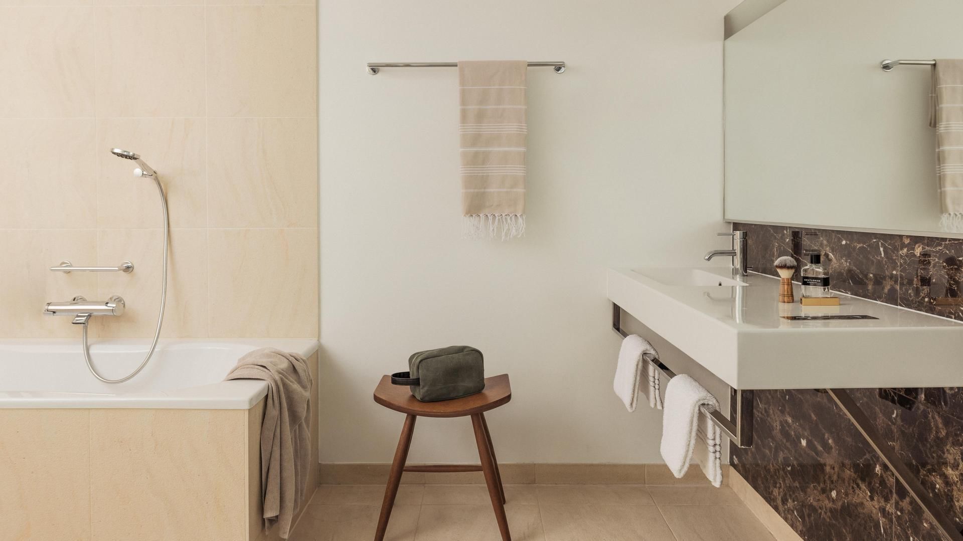 Modern bathroom with bathtub, showerhead, stool with bag, white sink, mirror, and towels.