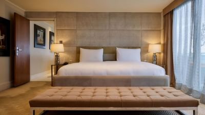 Spacious bedroom with king bed, tufted bench, textured wall panels, and floor-to-ceiling windows at The May Fair Hotel, London.
