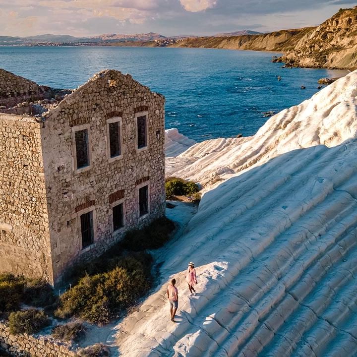 Ruiniertes Gebäude neben weißen Felsen und dem Meer mit zwei Menschen auf einem Pfad.