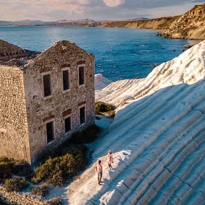 Ruiniertes Gebäude neben weißen Felsen und dem Meer mit zwei Menschen auf einem Pfad.