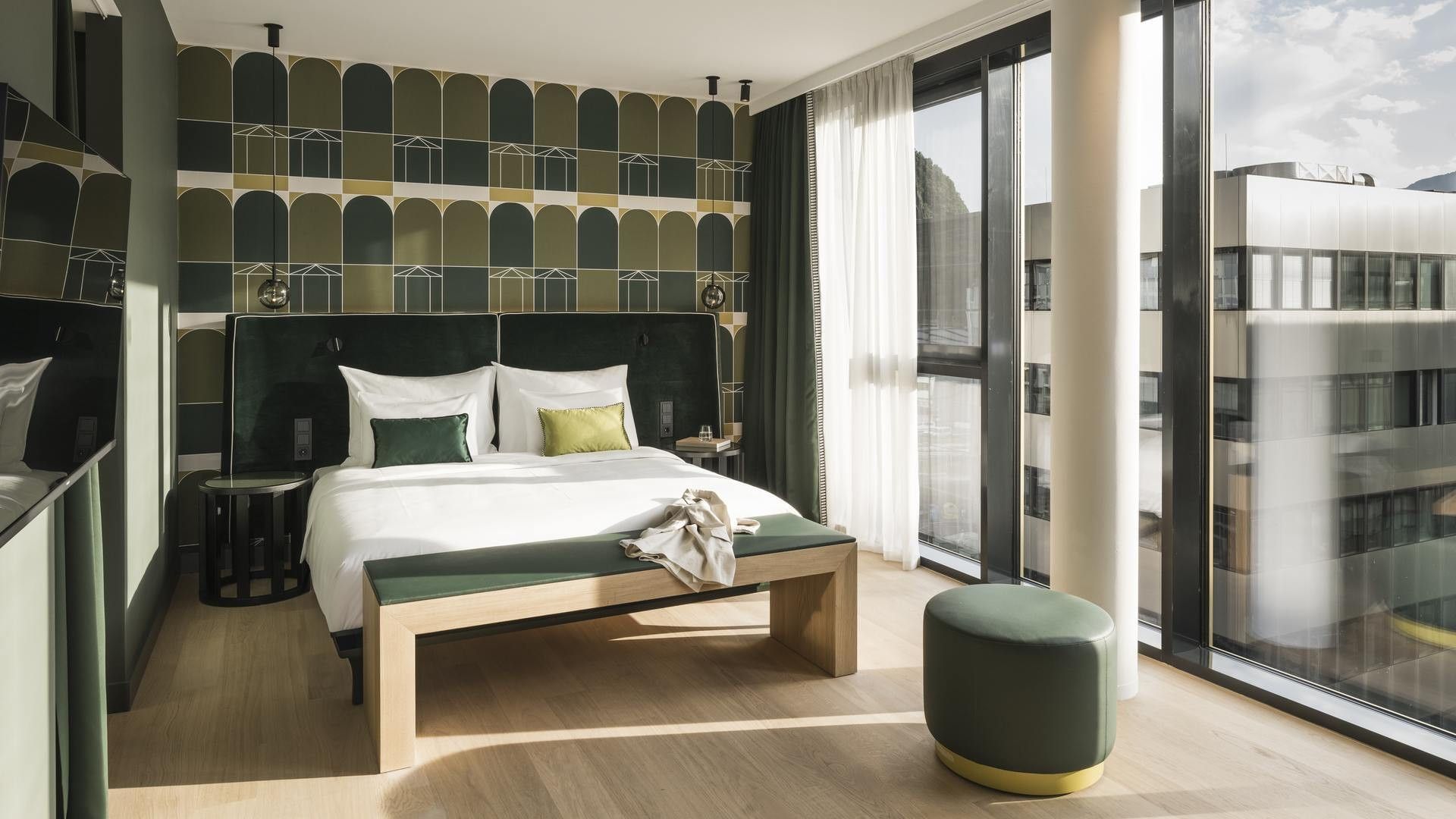Modernes Hotelzimmer mit großem Bett, Sitzbank, rundem Hocker und architektonischem Ausblick.