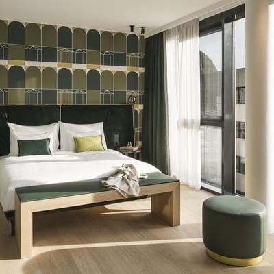 Modernes Hotelzimmer mit großem Bett, Sitzbank, rundem Hocker und architektonischem Ausblick.